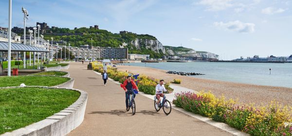 Two men cycling towards the camera on Dover Promenade with colourful flowers in beds, Dover Harbour, Dover Castle and the White Cliffs in the background