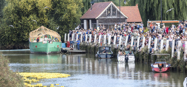 The River Stour and Sandwich Quay with crowds of people lining the riverbank to watch the annual duck race (yellow rubber ducks and boats in the water). Sandwich Medieval Centre in the background. 